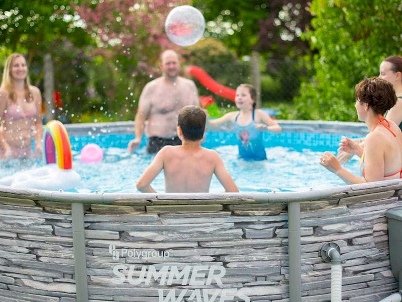 Familie im Summer Waves Aufstellpool, spielend mit Wasserbällen und aufblasbaren Spielsachen, umgeben von Gartenlandschaft